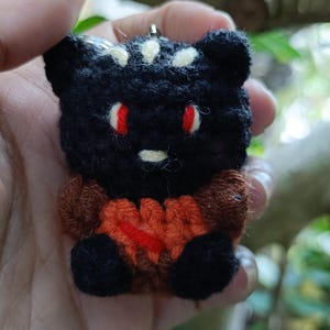 May include: A handmade black and brown crochet bear with red eyes and a white stripe on its head. The bear has a small orange heart on its chest and is a keychain.
