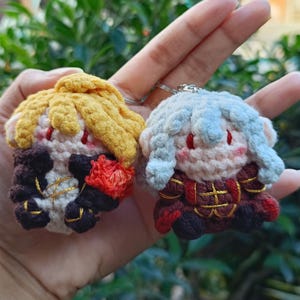 Può includere: Due piccoli portachiavi in peluche realizzati all'uncinetto a mano con personaggi stilizzati. Una figura ha i capelli gialli e un abito scuro con accenti rossi, mentre l'altra ha i capelli azzurri e un abito rosso scuro e nero.