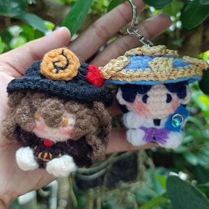 Könnte beinhalten: Zwei handgefertigte Häkel-Schlüsselanhänger mit Zeichentrickfiguren. Einer hat einen schwarzen Hut mit goldenem Emblem und rotem Akzent. Der andere hat einen blau-beigen Hut. Beide Schlüsselanhänger haben detaillierte Gesichtszüge.