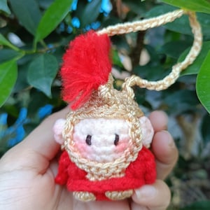Peut inclure: Une petite figurine crochetée portant un casque doré avec une crête rouge et une tunique rouge. La figurine a un visage clair avec des joues roses et des yeux sombres. Une boucle crochetée dorée est fixée au sommet du casque pour suspendre.