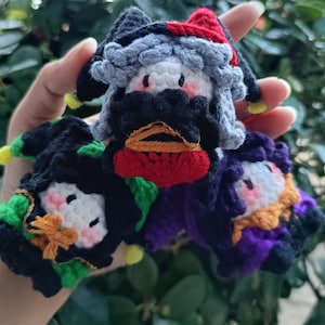 May include: Three small, handcrafted crochet plush figures featuring jester-style hats. One figure has a black and red hat with grey hair, one has a black and green hat with orange accents, and one has a black and purple hat with yellow details.