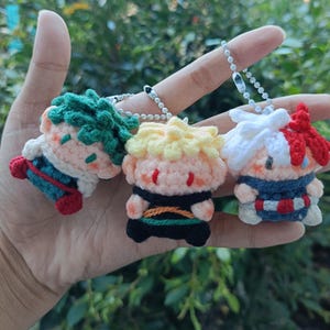 May include: Three handmade crochet character keychains. Each keychain features a different character with unique hair and clothing colors: green, yellow, and red. The keychains are attached to silver ball chains.
