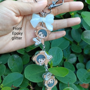 May include: A keychain featuring three cartoon character charms in bear costumes, connected by silver chains. The charms are in a descending order, with a white bow at the top and a silver clasp. The text "Front Epoxy glitter" is visible.