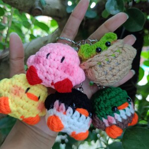 May include: A collection of five handmade crochet keychains. The keychains feature various animal designs, including a pink character, a yellow duck, a penguin, and two other creatures. Each keychain has a metal ring for attachment.