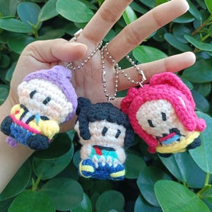 Puede incluir: Tres llaveros de muñecas de ganchillo hechos a mano. Cada llavero presenta una pequeña figura de muñeca con diferentes colores de pelo y atuendos. Las muñecas tienen ojos negros y están unidas a cadenas de bolas plateadas. El fondo es una exuberante planta verde.