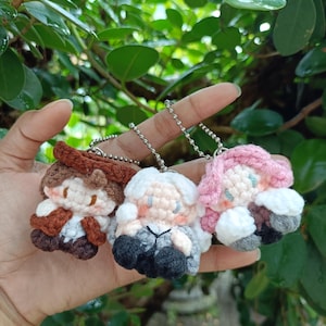 May include: Three crocheted doll keychains in a hand. The dolls have different hair colors: brown, white, and pink. Each doll has a unique outfit, with details like a hat and clothing. The keychains are attached to silver ball chains.