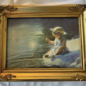 May include: A framed painting with a gold ornate frame. The artwork depicts a child wearing a hat and light blue dress, sitting near water and holding a stick. The painting has a soft, dreamy quality.