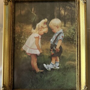 May include: A framed print featuring two young children in a forest setting. The boy wears a blue shirt and shorts, while the girl is in a white dress. The frame is ornate with a gold finish, adding a vintage aesthetic.