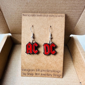 Könnte beinhalten: Ein Paar silberne Ohrringe mit roten AC/DC-Logo-Anhängern. Die Ohrringe werden auf einer braunen Karte mit dem Text "Please tag us @BLH. Jewellerydesign wearing our earrings!" und "Instagram: blh.jewellerydesign Etsy Shop: BLH Jewellery Design" präsentiert.