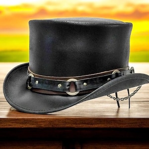 May include: A black leather top hat with a wide brim, brown leather band, and silver-colored metal accents. A silver chain is attached to the brim. The hat is displayed on a wooden surface.