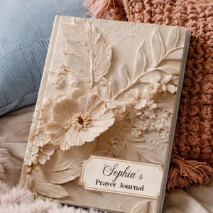 May include: A cream-coloured journal featuring a raised, three-dimensional floral relief design on the cover. A rectangular label at the bottom displays the text "Sophia's Prayer Journal" in a black serif font against a light background.