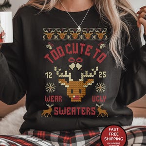 Puede incluir: Sudadera negra con un diseño festivo. El diseño incluye un reno, copos de nieve y el texto "TOO CUTE TO WEAR UGLY SWEATERS." La sudadera tiene cuello redondo y mangas largas. El diseño es en tonos rojos, marrones y blancos.