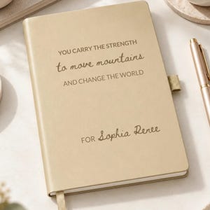 May include: A beige hardback journal featuring a gold pen holder loop and a ribbon bookmark. The cover displays the text "YOU CARRY THE STRENGTH to move mountains AND CHANGE THE WORLD" and "FOR Sophia Renee" printed in a dark brown, elegant font.