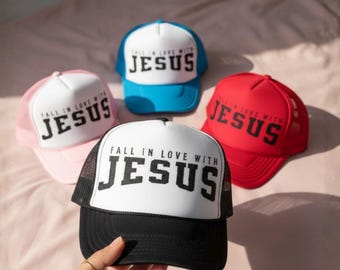 May include: A collection of trucker hats in various colors: pink, blue, red, and black. Each hat features a white front panel with the text "FALL IN LOVE WITH JESUS" in bold, black letters. The hats have mesh backs and curved brims.