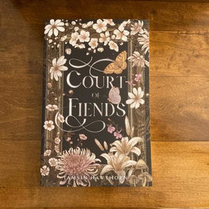 May include: A book titled Court of Fiends by Tamsin Hawthorn with a black cover featuring a decorative gold frame, white floral illustrations, a brown butterfly, and a small pink butterfly centered above the white serif text title.