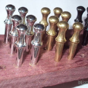 May include: A set of metal cribbage pegs in copper, silver, gold, and black finishes inserted into a purple-toned wooden board. The pegs feature a rounded top and a tapered body. The text www.enumerocribbageboards.com is visible at the bottom.