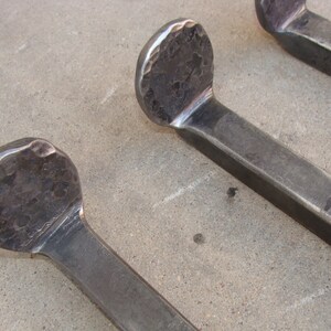 May include: Three hammered metal hooks with rounded ends. The hooks are made of a dark grey metal and have a rough, textured surface.