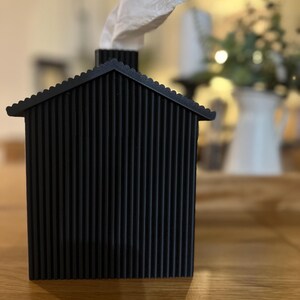 May include: A black, house-shaped tissue box cover with a corrugated design. White tissues emerge from the chimney. The tissue box is on a wooden table. The design is modern and minimalist.