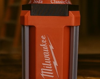 May include: An orange and black can cooler with the Milwaukee logo in white. The cooler is designed to hold a soda can, with the words "Soda Classic Cola" visible on the can.