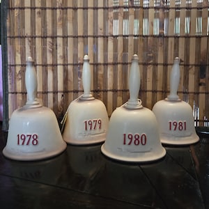 May include: Four cream ceramic bells with white handles, each inscribed with a year in red: 1978, 1979, 1980, and 1981. The bells are displayed on a dark surface against a bamboo backdrop.