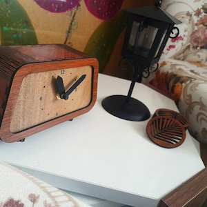 Puede incluir: Un reloj de madera con una esfera clara y manecillas negras se encuentra sobre una superficie blanca. El reloj es rectangular con un marco de madera marrón. Una linterna negra y dos posavasos de madera también están presentes.