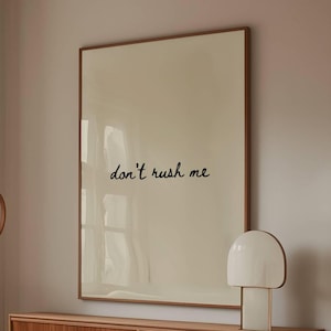 May include: A living room scene featuring a white, curved sofa, a wooden coffee table with a glass top, and a wooden cabinet. A large framed artwork with the text "don't rush me" hangs above the cabinet. A white lamp and a floor lamp are also present.