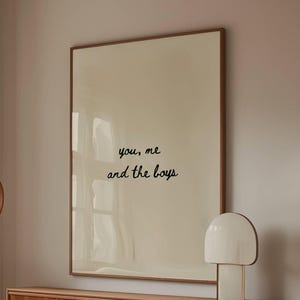 Może przedstawiać: Scena w salonie z kremową sofą, drewnianym stolikiem kawowym ze szklanym blatem i drewnianą szafką. Duży oprawiony wydruk z napisem "you, me and the boys" wisi nad szafką. Obecna jest również biała lampa i lampa podłogowa.