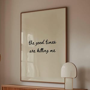 Może przedstawiać: Salon z białą, zakrzywioną sofą, drewnianym stolikiem kawowym ze szklanym blatem i drewnianą szafką. Duży oprawiony wydruk na ścianie głosi "the good times are killing me". Widoczna jest również biała lampa i lampa podłogowa.