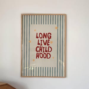 以下が含まれることがあります： 「LONG LIVE CHILDHOOD」の赤い文字と黄色の星のアクセントが入った額入りのプリント。プリントはクリーム色の背景に青と白のストライプのボーダーがあります。フレームは明るい茶色です。