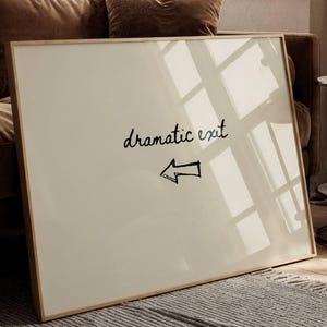 Puede incluir: Una impresión artística enmarcada con las palabras "dramatic exit" en cursiva y una flecha que apunta hacia la izquierda. La impresión tiene un fondo color crema y un marco de madera delgado. La impresión está apoyada contra un sofá marrón.