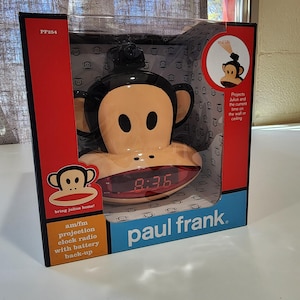 May include: A Paul Frank Julius the monkey AM/FM projection clock radio with battery back-up. The clock displays 8:36 in red digits. The packaging is red, black, and blue, with the Paul Frank logo and the text "bring julius home!"
