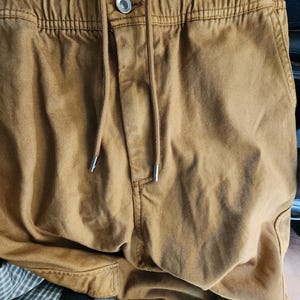 May include: Close-up of tan-colored drawstring pants. The pants have a button closure and a drawstring with silver-colored metal tips. The fabric appears to be a sturdy cotton or canvas material, with visible stitching details.