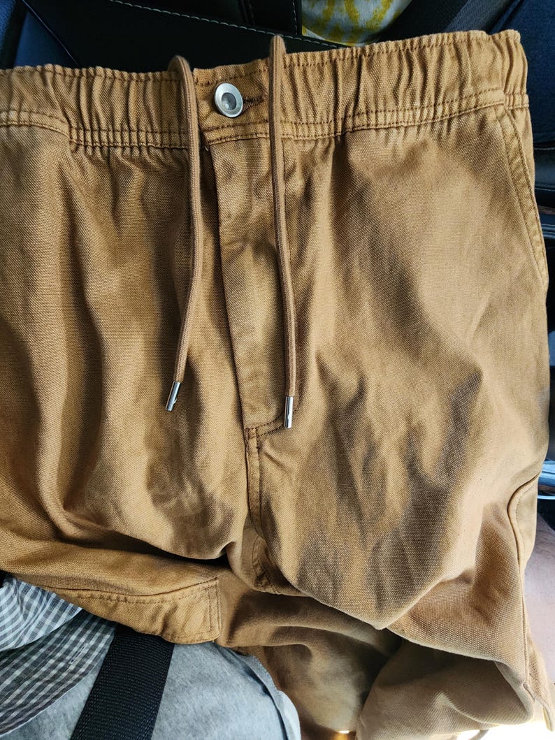 May include: Close-up of tan-colored drawstring pants. The pants have a button closure and a drawstring with silver-colored metal tips. The fabric appears to be a sturdy cotton or canvas material, with visible stitching details.