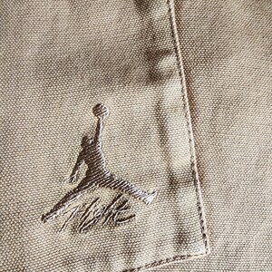 May include: Close-up of a tan fabric with a stitched pocket and the embroidered Jumpman logo in a darker tan color. The logo features a silhouette of a basketball player in mid-air, with the Nike signature below.