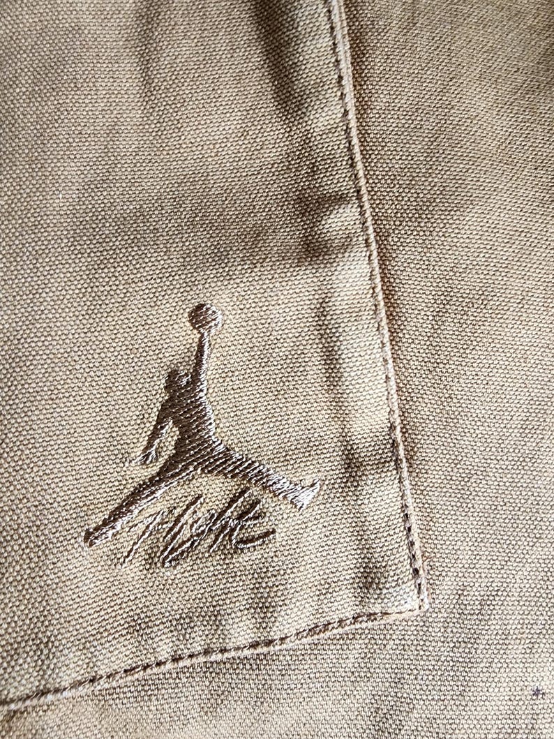 May include: Close-up of a tan fabric with a stitched pocket and the embroidered Jumpman logo in a darker tan color. The logo features a silhouette of a basketball player in mid-air, with the Nike signature below.