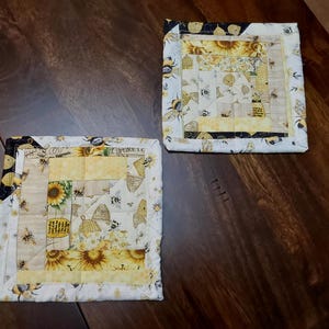 May include: Two square quilted pot holders with a bee and sunflower theme. The pot holders are white with yellow, black, and brown accents. The fabric features a repeating pattern of bees, honeycombs, and sunflowers.