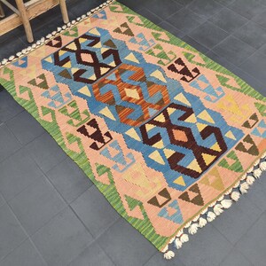 May include: Rectangular woven rug with a geometric pattern. The rug features a central design in blue, orange, and brown, surrounded by pink and green borders. White tassels adorn the edges. The rug is approximately 3 x 5 feet.
