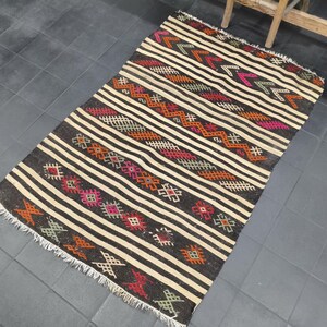 May include: A rectangular, hand-woven rug with a dark brown base and cream-colored stripes. The rug features geometric patterns in shades of red, orange, pink, and green. The edges are finished with a fringe.