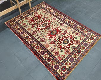 Puede incluir: Alfombra rectangular con un diseño geométrico central en rojo, crema y azul. La alfombra presenta un borde con patrones florales y geométricos repetidos en rojo, morado y crema. La alfombra está colocada sobre un suelo de baldosas grises.