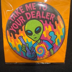 May include: A round button with a green alien holding a purple bong, wearing a tie-dye shirt. The button has the text "TAKE ME TO YOUR DEALER" in pink and white, with a colorful psychedelic background.