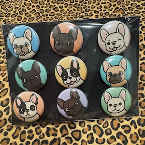 May include: Nine round pinback buttons featuring French Bulldog illustrations. Each button has a different colored background, including blue, pink, and green. The dog illustrations vary in color and expression, with some smiling and others with a neutral expression.