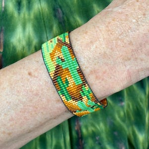 May include: A woven bracelet with a geometric pattern in shades of green, brown, and yellow. The bracelet is worn on a wrist, showcasing the intricate beadwork and color combination. The bracelet is approximately 1.5 inches wide.