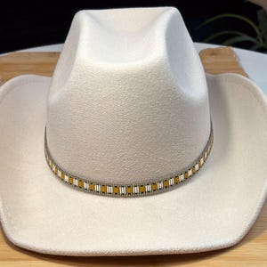 May include: A cream-colored cowboy hat with a decorative band featuring gold and silver rectangular accents. The hat has a classic, wide brim and a rounded crown. The hat is made of a soft, felt-like material.