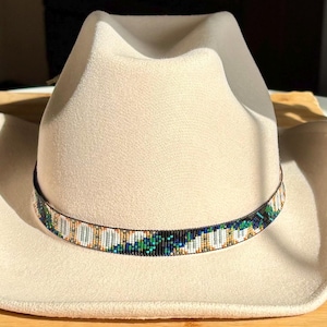 May include: A cream-colored cowboy hat with a decorative beaded band. The band features a pattern of green, white, and gold beads. The hat is made of a soft material and has a classic cowboy hat shape.