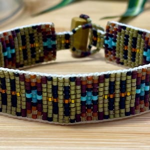 May include: A beaded bracelet with a geometric pattern. The bracelet features a cream-colored base with a design of olive green, black, burgundy, orange, and turquoise beads. The clasp is made of wood.