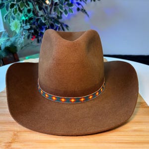 May include: A brown felt cowboy hat featuring a pinched crown and a wide brim. The hat is accented with a narrow, decorative band made of small, colorful beads arranged in a repeating geometric pattern of orange diamonds on a teal background.