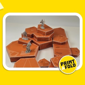 Pode incluir: Um conjunto de peças de terreno de cartão castanho para jogos de mesa. As peças têm a forma de rochas hexagonais e estão dispostas numa formação em camadas. As peças são concebidas para serem impressas e dobradas, como indica o texto "PRINT & FOLD" num círculo amarelo.