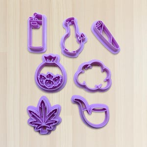 Puede incluir: Un conjunto de siete cortadores de galletas de plástico morado con formas variadas: un encendedor, un bong, un objeto enrollado, una bolsa de flores, una nube, una hoja de marihuana y una pipa. Dispuestos sobre una superficie de madera clara.