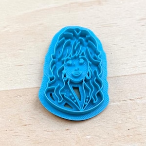 May include: A bright blue cookie cutter featuring a raised, molded design of a person with long, wavy hair and a collared jacket. The tool has a flat base and a detailed, embossed face and hair texture for creating impressions in dough or clay.