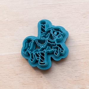 May include: A teal plastic cookie cutter shaped like a carousel horse. The design features a horse with a saddle, a pole, and a flowing mane, with a raised outline that creates a detailed impression when pressed into dough or other soft materials.